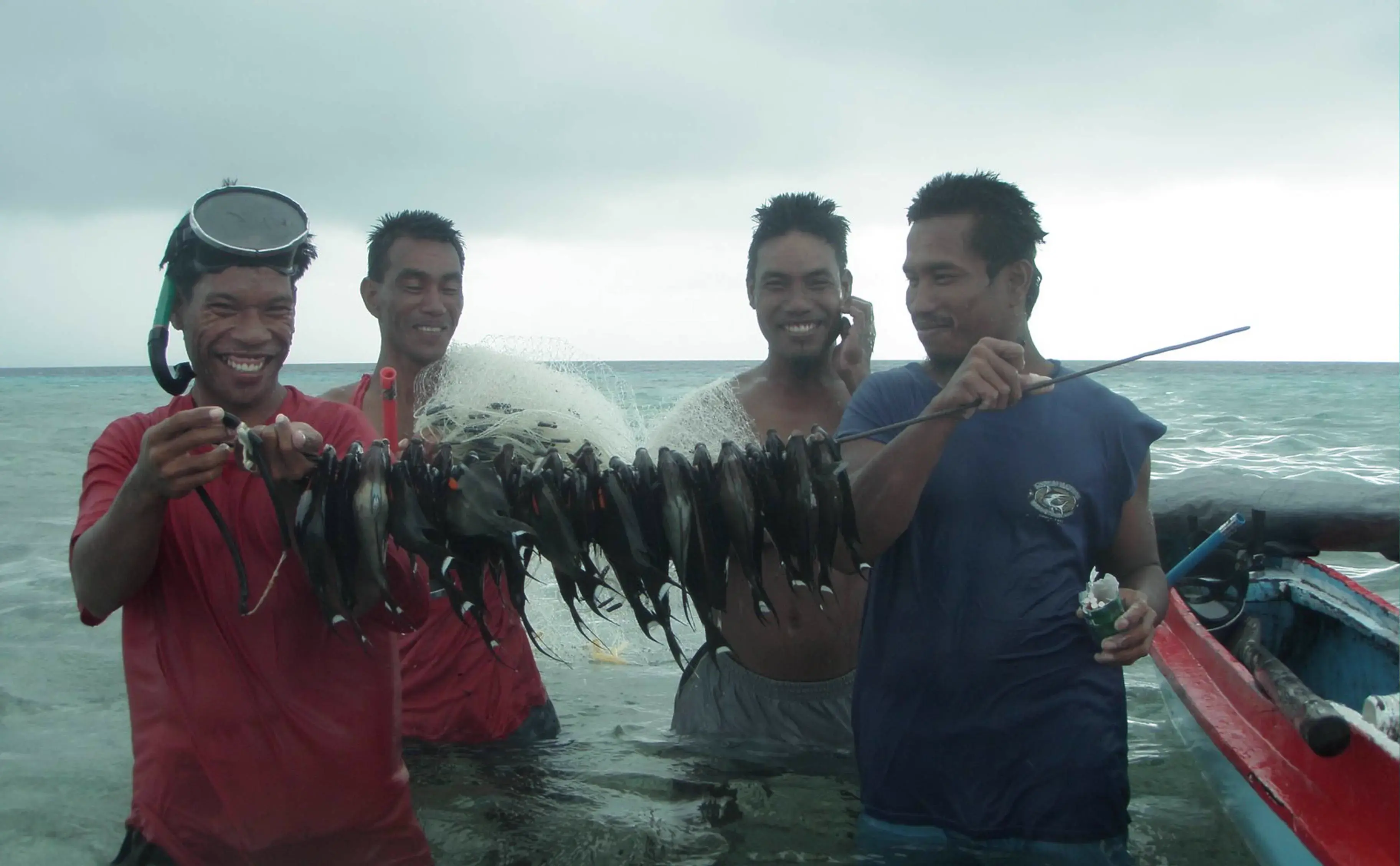 Four local fishermen smiling while holding a large net full of fish on a boat.